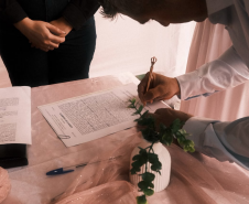 Homem, assinando documento em uma mesa de toalha branca. Ele está usando um terno e perto de si tem um pequeno vaso com uma rosa
