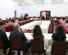 Vemos todos os participantes do encontro. A foto foi tirada atrás da mesa principal dos palestrantes, então vemos esses de costas e os participantes sentados em largas mesas laterais e uma frontal. 