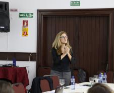 A Assistente Social do MPPR, Clarice Metzner, está de pé atrás da mesa principal para os palestrantes falando. 