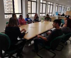 Mesa da reunião, os representantes estão sentados em volta da mesa. De costas, no início da foto, está presente a dra. Olenka Lins.