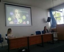 A imagem consiste da Dra. Luciana Tramujas, coordenadora da defensoria da Casa da Mulher Brasileira, e da Dra. Eliana Lopes, Coordenadora do Pró-mulher, sentadas em uma mesa (cada uma em uma ponta) e ao fundo um tela com uma apresentação sobre violência e gênero. 