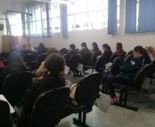 A imagem foi tirada no fundo da sala, focando nos ouvintes. Vemos esses de costas e sentados. 