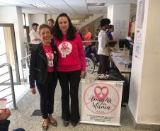 As duas palestrantes encontram-se juntas, no CAM Curitiba, após o termino da palestra. Ambas estão sorrindo para a foto e há um cartaz da Associação Amigas da Mama ao seu lado. 