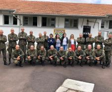 Vemos os soldados, alunos do curso, em fileiras - alguns abaixados e outros de pé - posando para foto. A equipe da DPPR da Casa da Mulher Brasileira se encontra em pé, no meio dos soldados, 