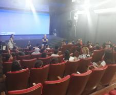 Na imagem vemos o encerramento do projeto no auditório da UNICENTRO. Temos os alunos sentados nas poltronas e as professoras em frente ao palco e telão. 