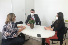 A foto mostra três pessoas, um homem e duas mulheres, sentados à mesa, usando máscaras