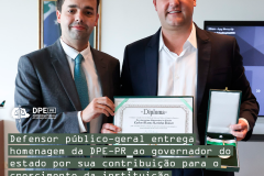 Imagem com a foto do Defensor Público-Geral, André Giamberardino, entregando ao governador do estado do Paraná, Carlos Massa Ratinho Junior, um certificado e uma medalha de homenagem pela colaboração com o crescimento da Defensoria.  Os dois são homens caucasianos altos e com cabelos castanhos. Ambos usam terno e gravata. Alinhado à esquerda, o título da matéria, tarjado em verde e, acima, o logo da Defensoria em branco.