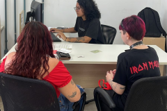 Imagem que mostra duas pessoas sendo atendidas no mutirão em Ponta Grossa. Elas estão de costas e entre elas, é possível ver a pessoa que está fazendo o atendimento, sentada atrás da mesa e olhando para a tela do computador.