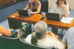 Imagem de uma foto de um atendimento presencial, duas servidoras públicas estão conferindo informações em seus notebooks enquanto três usuários da Defensoria Pública aguardam. Na parte central inferior da imagem, está o título da matéria.