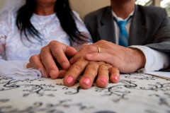 casal com as mãos sobre a mesa mostrando as alianças. eles usam trajes de terno e gravata e vestido de noiva.