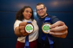 Imagem colorida horizontal em que se vê no primeiro plano as medalhas da DPE-PR e ao fundo os servidores premiados. 