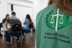 Imagem horizontal colorida em que se vê colete da defensoria em primeiro plano e ao fundo desfocados assistidos recebendo atendimento durante mutirão.