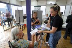 Imagem colorida horizontal em que se vê uma senhora idosa recebendo um panfleto de uma moça com informações sobre os serviços da DPE-PR durante o evento Assembleia Itinerante realizado em Londrina.