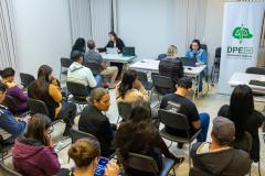 Imagem colorida horizontal em que se vê uma sala cheia de pessoas e ao fundo a equipe da DPE-PR realizando atendimento. 