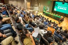 Imagem mostra o auditório quase cheio para a posse dos servidores, no palco ocorre uma apresentação músicas de violão e voz. Ao fundo mesa para as autoridades e o telão com logo da DPE-PR. 