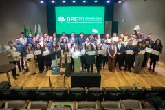 Grupo de Servidores empossados posam juntos para foto no palco de auditório. Ao fundo tela de Led mostra a logo da instituição.  