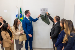 A imagem captura o momento do descerramento da placa inaugural. O Defensor Público-Geral, à esquerda, e o vice-prefeito de Foz do Iguaçu, à direita, seguram juntos um tecido verde-escuro, revelando a placa fixada na parede. Ao redor deles, um grupo de convidados, majoritariamente mulheres, observa a cena com sorrisos e expressões de satisfação.