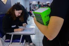 Imagem mostra braço de candidata segurando livro em close no primeiro plano enquanto fiscal confere seus documentos para ingressar na sala para do concurso. 