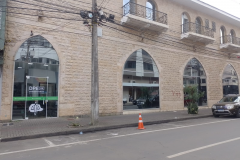 Imagem que mostra o prédio da nova sede da DPE em União da Vitória. O prédio possui fachada do andar térreo revestida em pedras de cor clara e marcada por grandes janelas e uma porta em formato de arco ogival. O andar superior também apresenta janelas em arco com sacadas e grades de ferro. À esquerda, uma porta de vidro exibe o logotipo e o nome da instituição. Através do vidro, é possível ver parte do interior da recepção.
