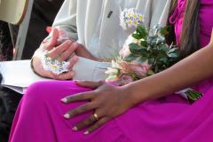Uma imagem focada nas mãos de um casal. A noiva, usando um vestido vibrante de cor magenta, tem uma aliança dourada no dedo e as unhas pintadas. Sua mão repousa sobre a perna, próxima a um buquê. As mãos do noivo, que veste uma camisa clara, estão unidas e adornadas com um pequeno arranjo de margaridinhas amarrado em seu pulso.