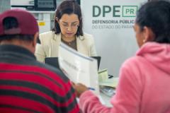 Assistidos aparecem de costas desfocados entregando documentos à servidora da DPE-PR em Mutirãop de Conciliação. 