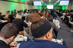 Homem negro com tranças e homem branco tomando notas em caderno, ambos de costas em primeiro plano, e ao fundo se vê a sala lotada de defensores de too o país na abertura di Conajuri.