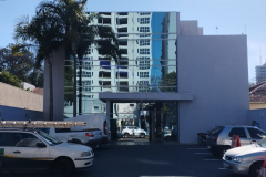 Imagem que mostra a fachada da sede da Defensoria Pública em Cianorte, em um dia claro e ensolarado. O prédio é uma construção moderna de dois andares, com uma grande seção central de vidro espelhado. As laterais do prédio são de alvenaria, pintadas em um tom de cinza-claro. Na frente do edifício há um estacionamento com alguns carros parados. À esquerda do prédio, uma palmeira alta se destaca contra o céu.