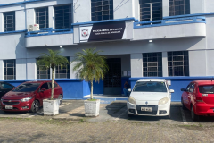 A foto mostra a fachada da Cadeia Pública de Paranaguá, um prédio de dois andares, pintado em tons de azul claro e escuro, sob um céu limpo. Em frente, há um estacionamento com carros.