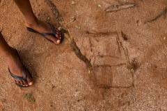 Imagem mostra detalhe dos pés de uma criança de chinelo usando um galho de madeira para desenhar um cubo na terra avermelhada. 
