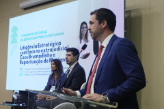 Um homem de terno azul e gravata vermelha discursa em um púlpito. Atrás dele, duas pessoas estão sentadas no palco, e um telão exibe o título da palestra: "Litigância Estratégica com foco no extrajudicial".