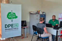 Visão mais ampla da sala com um banner vertical da DPE-PR em destaque à esquerda. Ao fundo, o funcionário de verde usa o notebook enquanto atende uma mulher de blusa cinza sentada à sua frente.