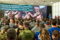 Imagem mostra o salão de atos do Palácio Iguaçu lotado com oficiais das forças de segurança. Ao fundo letreiro sobre o lançamento do NUSEG e o DPG Matheus Munhoz discursando.  