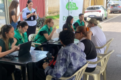 Em uma tenda de atendimento na rua a equipe da Defensoria Pública conversa e orienta as pessoas sentadas à frente das mesas. Ao fundo, destaca-se um banner branco e verde da DPE-PR.