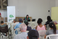 Uma mulher de colete verde trabalha em um notebook ao fundo da sala, ao lado de um banner da DPE-PR. À frente dela, diversas pessoas aguardam sentadas de costas, incluindo uma pessoa em cadeira de rodas.