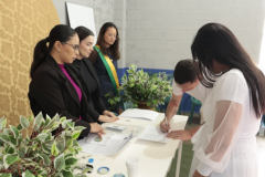 Um homem de camiseta branca assina um documento sobre uma mesa, ao lado de uma mulher de vestido branco. Atrás da mesa, três mulheres observam atentamente, sendo que uma delas usa uma faixa transversal com as cores da bandeira do Brasil.