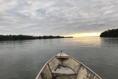 A foto foi tirada dentro de um barco simples de pescador. Vemos a  parte frontal do barco, seu bico, o barco está no rio e ao fundo vê-se o pôr do sol. 