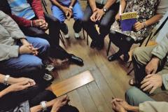 A foto foi tirada sob uma perspectiva de cima; Vemos um grupo de pessoas sentadas em roda, todas muito juntas, simulando uma reunião de AA. 