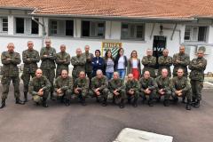Vemos os soldados, alunos do curso, em fileiras - alguns abaixados e outros de pé - posando para foto. A equipe da DPPR da Casa da Mulher Brasileira se encontra em pé, no meio dos soldados, 