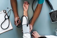 Imagem de um médico, onde esse está medindo a pressão do paciente.
 A foto encontra-se em uma perceptiva observada de cima, aparecendo apenas os braços de ambos. 
