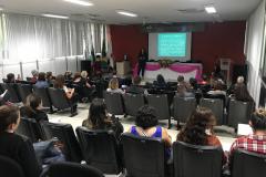 Imagem tirada durante a palestra do funda da sala. Vemos as costas dos ouvintes e a palestrante a frente. 