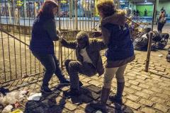 A imagem, uma foto ilustrativa, registra um morador de rua, idoso, sendo atendido, à noite, por duas assistentes sociais.