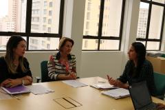 Descrição da imagem: Na foto estão: da esquerda para a direita, a coordenadora de saúde mental da Secretaria Municipal da Saúde (SMS), Flávia Adachi, a secretária municipal de saúde de Curitiba, Marcia Cecilia Huçulak e a coordenadora do Núcleo da Cidadania e Direitos Humanos (NUCIDH) da Defensoria Pública do Paraná, Cinthia Azevedo Santos. Ambas estão sentadas com papéis sobre a mesa. Ao fundo aparecem três grandes janelas, em uma parede branca, com prédios aparecendo.