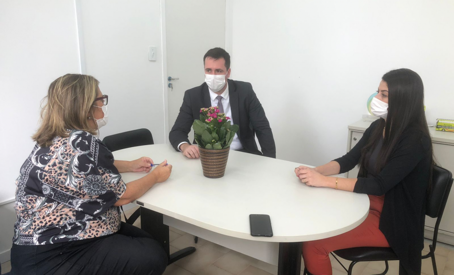 A foto mostra três pessoas, um homem e duas mulheres, sentados à mesa, usando máscaras