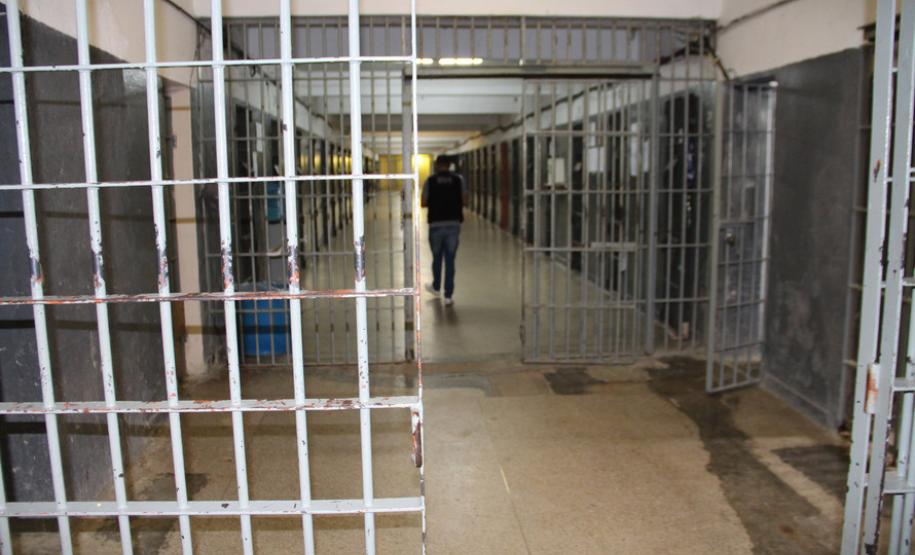 Foto mostra o interior de uma prisão, com um agente penitenciário de costas e um cela com a porta aberta