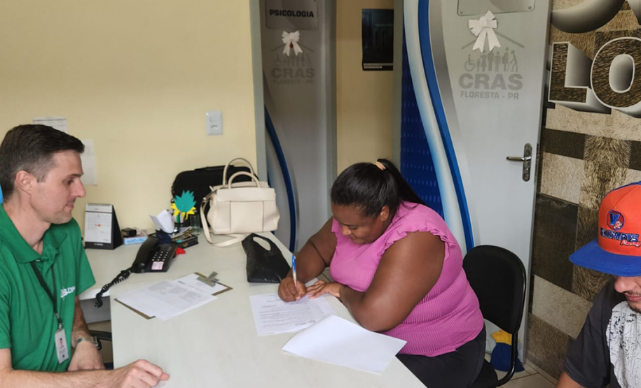Foto de atendimento com um servidor da defensoria de pele branca camiseta verde com o logo da instituição e calça jeans, acompanhado de dois usuários assinando um documento, sendo a mulher negra com uma camisa cor de rosa e calça preta e o homem branco com calça jeans, camisa preta e boné azul com detalhes em vermelho.