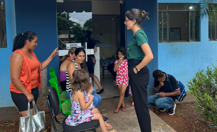 Foto de uma servidora branca com calça preta e camiseta da instituição na cor verde, conversando com usuárias da defensoria que esperam sentadas em cadeiras do lado de fora do prédio do CRAS. O prédio é de cor azul, em frente estão três cadeiras verdes e uma preta, onde estão sentadas duas crianças acompanhadas de suas mães, sendo elas uma mulher negra e duas mulheres brancas. Mais ao lado, uma criança branca aguarda ao lado da porta e, agachado também ao lado da porta, um homem mexe no celular.