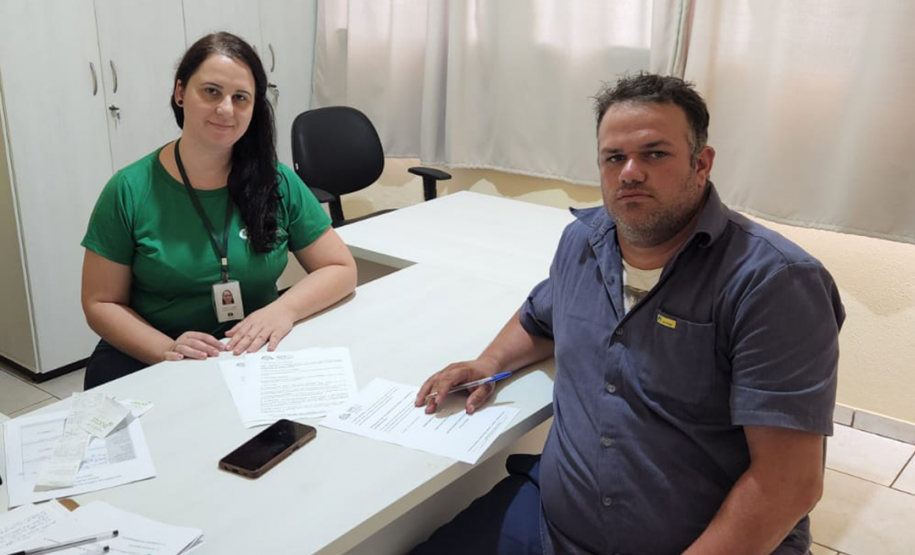Imagem de uma servidora da defensoria com pele branca e camiseta da instituição na cor verde, com os braços apoiados em uma mesa branca. Ela atende um usuário branco com camiseta e calça em tons de azul. Ele segura uma caneta para assinar alguns papeis que estão sobre a mesa.