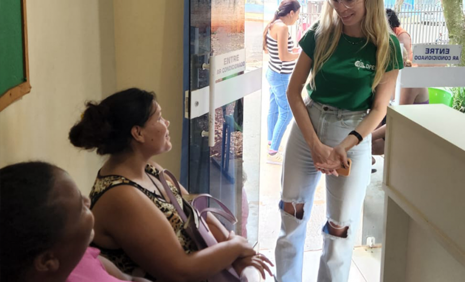 Imagem de duas mulheres negras usuárias da defensoria sentadas na parte interna do prédio do CRAS sendo atendidas por uma servidora branca que usa a camiseta da instituição e calça jeans.
