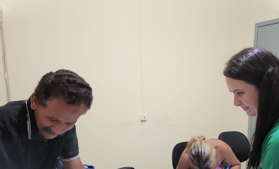 Imagem de uma servidora da Defensoria com a camisa da instituição em verde entregando documento para um homem branco e, ao fundo, uma mullher branca assina alguns papeis em uma mesa branca.