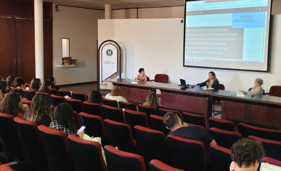 Imagem que mostra uma das palestras do evento. Três mulheres fazem uma apresentação, sentadas atrás de uma mesa, para uma plateia formada por várias pessoas que as escutam com atenção.